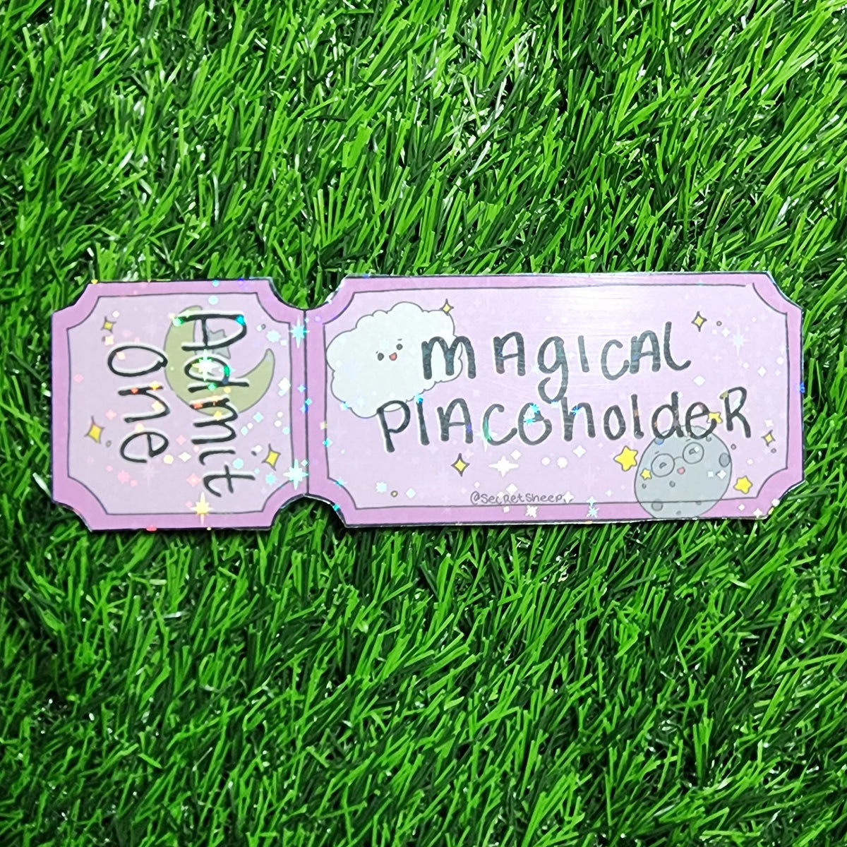A pink double-sided card or plaque reading 'Admit One' and 'Magical Placeholder' with small moon, cloud, and star illustrations, placed on bright green artificial grass.
